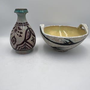 May include: Two ceramic items are shown: a small vase and a bowl. The vase is off-white with dark brown floral patterns and a turquoise rim. The bowl has a cream interior with black and white accents on the exterior and two handles.