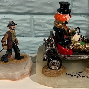 May include: Two decorative figurines. One features a clown pulling a red wagon with puppies. The other shows a clown in a go-kart. Both figurines are on a white base with a sand-like texture.
