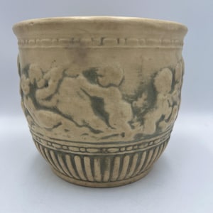 Puede incluir: Una maceta de cerámica beige con un diseño en relieve detallado. La maceta presenta una banda de figuras de querubines alrededor del centro y ranuras verticales en la base. El borde tiene un borde decorativo. La estética general es clásica.