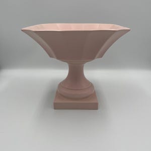 Puede incluir: Un cuenco rosa pálido sobre pedestal, con un borde ancho y festoneado. Presenta una columna acanalada y una base cuadrada. El diseño simétrico sugiere que es de cerámica o un material similar. El cuenco es probablemente decorativo.