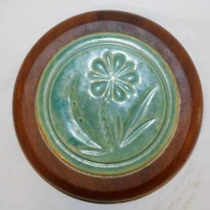 May include: Circular wooden trivet with a teal ceramic centre. The ceramic insert has a floral design, with a central flower and leaves. The trivet is a warm brown colour and is likely used for hot dishes.