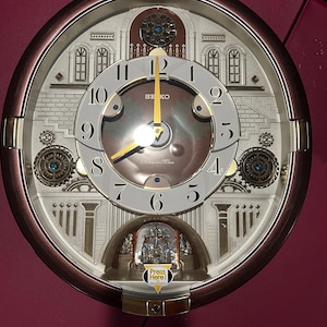 May include: A round Seiko wall clock with a metallic bronze frame and a white face featuring black Arabic numerals. The clock includes gold hands, decorative gear-like accents with blue crystals, and a small sticker that reads Press Here.