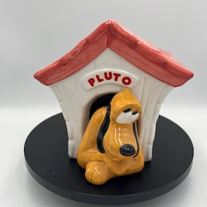 May include: A ceramic figurine of Pluto the dog in his doghouse. The doghouse is white with a red roof and the name "PLUTO" in red letters. Pluto is yellow with black ears and a black nose. The figurine sits on a black circular base.