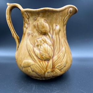 May include: A tan ceramic pitcher with a handle and a floral design. The pitcher has a bulbous body and a flared rim. The floral design features a large tulip and several smaller buds. The pitcher is glazed with a speckled finish.