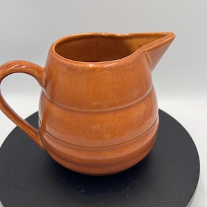 Puede incluir: Una jarra de cerámica esmaltada en un tono naranja cálido, con un cuerpo redondeado y abombado con tres bandas horizontales en relieve que rodean el exterior. El recipiente incluye un asa curva en el lateral y un pico vertedor puntiagudo.