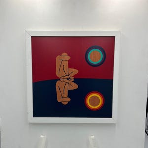May include: Abstract art piece in a white frame. The artwork features a figure in a seated position, with a reflection below. The background is divided into red and navy blue sections, with two circular designs in the upper right and lower right corners.