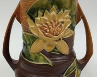 May include: A brown and tan ceramic vase with a water lily design. The vase has two handles and a textured surface. The water lily is yellow and green, with detailed petals and leaves. The vase is a decorative item.