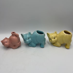 May include: Three ceramic cat figurines in pastel colors. The first is pink, the second is light blue, and the third is yellow. Each cat has a different design, with the blue and yellow cats having openings on their backs.
