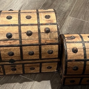 May include: Two wooden storage chests with a rounded, domed lid design. Each chest features a light brown wood grain finish, reinforced with dark metal grid straps and decorative circular metal studs at each intersection of the grid pattern.