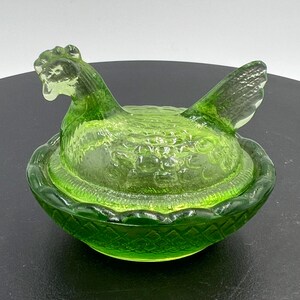 Puede incluir: Un plato de cristal verde lima translúcido con forma de gallina sobre un nido. La tapa tiene forma de gallina sentada con detalles de plumas texturizados, mientras que la base presenta un patrón de cesta tejida con un borde festoneado.