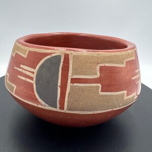 May include: A hand-painted, red ceramic bowl with geometric designs in tan, black, and white. The bowl has a rounded shape with a wide opening. The design includes a half-circle and rectangular shapes.