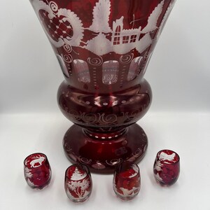 May include: A ruby red glass vase and four matching shot glasses. The vase features a detailed etched design of a cityscape. The shot glasses have etched forest scenes. The set is a vintage style.