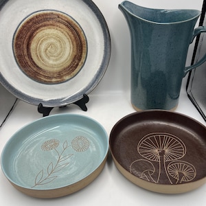 May include: A collection of handmade ceramic tableware. Includes a large plate with a spiral design, a tall blue pitcher, and two small bowls. One bowl is light blue with a floral design, and the other is brown with a mushroom design.