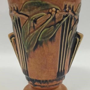 May include: A brown ceramic vase with a pedestal base and flared top. The vase features a relief design of stylized leaves and flowers in shades of green and brown. Vertical black lines add detail.