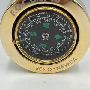 Puede incluir: Una brújula de latón con una esfera negra que muestra marcas direccionales y una aguja. La brújula tiene el texto "F.O.C.U.S. '98" y "RENO NEVADA" grabado. Es una herramienta de navegación.