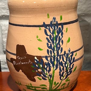 May include: A beige ceramic jar with a blue and green floral design. The jar features a brown outline of Texas with the words "Texas Bluebonnets" in white. Two dark blue stripes encircle the jar.