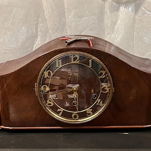 Op de afbeelding: Een vintage houten mantelklok met een gebogen bovenkant en een ronde wijzerplaat. De wijzerplaat heeft goudkleurige cijfers en wijzers. De klok is bruin met decoratieve gravures aan de zijkanten.