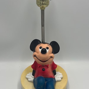May include: A vintage Mickey Mouse lamp. The lamp features a smiling Mickey Mouse figure in a red shirt, blue pants, and yellow shoes. The lamp has a brass-colored socket and a silver pole. The base is a light yellow color.
