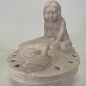 May include: A vintage ceramic figurine in a light beige color. The figurine depicts a seated child with a duck and a base with several holes. The ceramic has a cracked glaze finish.