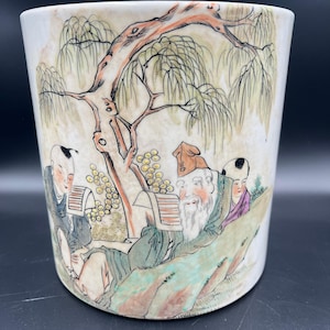 May include: A cylindrical ceramic vase with a hand-painted scene of figures under a tree. The artwork features a bearded man, other figures, and a weeping willow tree in muted colors. The vase has a cream-colored background.