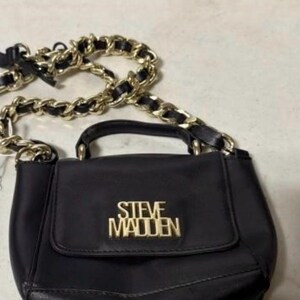 May include: A black handbag featuring a top handle and a flap closure with the gold-toned text STEVE MADDEN centered on the front. The bag includes a long shoulder strap made of interwoven black material and gold-toned metal chain links.