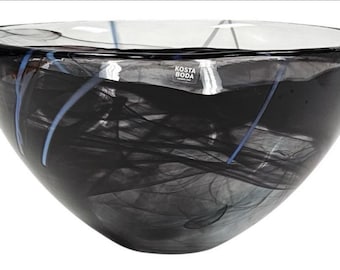 Può includere: Una ciotola di vetro trasparente con un design astratto nero e grigio. La ciotola ha una forma ampia e arrotondata con un bordo leggermente svasato. Strisce blu e bianche accentuano l'interno scuro. Il marchio "KOSTA BODA" è visibile.