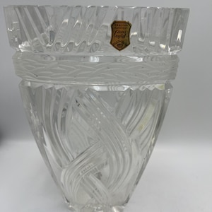 May include: Clear crystal vase with a wide, textured rim and a decorative band. The vase features a swirling, etched design and a label that reads "Genuine Hand Cut Lead Crystal".