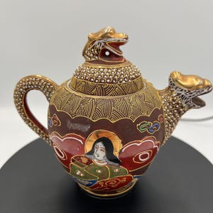 May include: A decorative brown ceramic teapot with gold accents and a dragon-shaped spout and lid. The teapot features a painted image of a woman with a halo and intricate gold detailing. The handle and lid are adorned with a textured pattern.