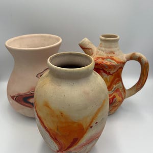 May include: Three ceramic vases with a marbled design. The vases are in shades of beige, orange, and red. One vase is a pitcher with a handle and spout. The other two are rounded with wide openings.