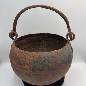 Peut inclure: Un chaudron rond en métal lourd doté d'une anse arquée épaisse fixée par des boucles circulaires de chaque côté. La surface extérieure présente une oxydation importante, avec une texture rouillée brun foncé et des motifs géométriques gravés.