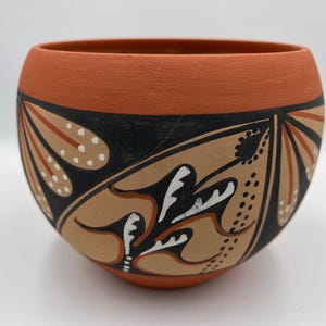Puede incluir: Un cuenco de cerámica de color terracota con forma redondeada. El cuenco presenta un diseño geométrico negro y beige con detalles blancos. El borde del cuenco es de color terracota liso.