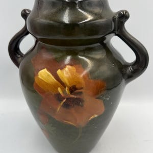 Puede incluir: Un jarrón de cerámica verde oscuro con dos asas laterales y un cuello ancho. El cuerpo del jarrón presenta un diseño floral pintado a mano en tonos naranja y amarillo con pinceladas visibles. La superficie tiene un acabado brillante.