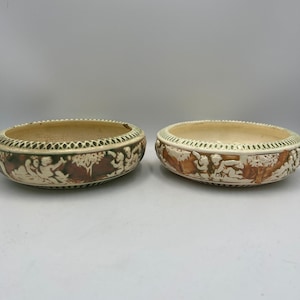 May include: Two cream-colored ceramic bowls with a decorative border and relief scenes. The bowls have a light brown and green color scheme. The relief scenes depict figures and trees. The bowls are oval-shaped.