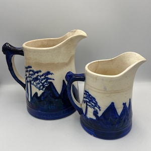 May include: Two ceramic pitchers with a cream-colored exterior and blue mountain and tree designs. The pitchers have blue handles and bases. The larger pitcher is approximately 9 inches tall, and the smaller pitcher is about 6 inches tall.