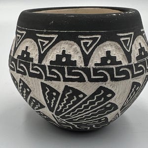 Può includere: Un piccolo vaso rotondo in ceramica bianca e nera con intricati motivi geometrici. Il design presenta forme e linee ripetute, creando un aspetto strutturato. Il bordo è nero e l'interno è di un colore più chiaro.