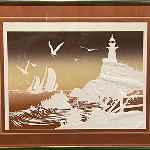 May include: Framed artwork depicting a coastal scene with a lighthouse, sailboats, and seagulls. The artwork features a white lighthouse on a cliff, sailboats on the water, and seagulls in flight against a gradient brown and yellow sky. The artwork is framed in a gold and brown frame.
