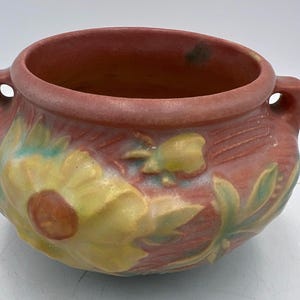 May include: A ceramic bowl with a reddish-brown exterior and interior. The bowl features a floral design in yellow, green, and brown. It has two small handles on either side. The bowl is likely used for decorative purposes or as a planter.