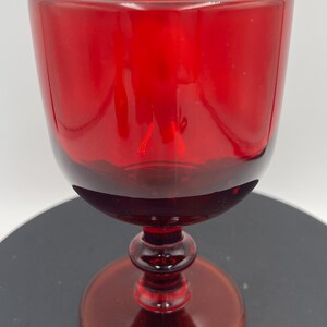 Peut inclure: Un gobelet en verre rouge uni doté d'un bol arrondi et lisse, d'une tige courte avec un bouton décoratif central et d'une base circulaire. Le verre est translucide, laissant passer la lumière à travers la couleur cramoisie profonde.
