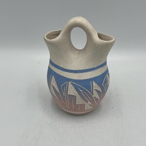 May include: A cream-colored pottery vase with a handle. The vase features a blue and pink geometric design. The top has a blue stripe. The vase is likely handmade and has a rustic aesthetic.