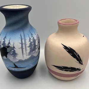 May include: Two ceramic vases. One is navy blue with a landscape scene of trees, water, and an orca whale. The other is a light tan color with black feather designs and a pink band around the base and rim.