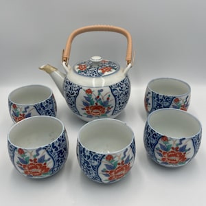 May include: A ceramic tea set featuring a teapot with a woven handle and six matching cups. The teapot and cups are white with blue, red, and green floral designs. The set is designed for serving tea.