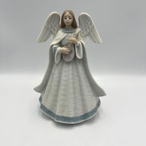 May include: A ceramic angel figurine with white wings and a flowing white gown with a blue trim. The angel has brown hair and is holding a musical instrument. The figurine is a decorative item.