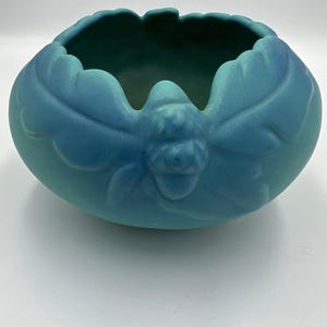 May include: A teal-colored ceramic bowl with a leaf-like design around the exterior. The bowl has a rounded shape with a scalloped rim and a detailed acorn-like emblem on the front. The interior is a darker shade of teal.