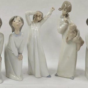 Può includere: Una collezione di cinque statuette in porcellana raffiguranti giovani figure in camicie da notte bianche. Le figure hanno pose diverse, tra cui allungarsi, coprirsi gli occhi e tenere in braccio una bambola. Le statuette hanno i capelli grigi e sono su sfondo bianco.