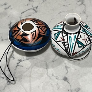 Könnte beinhalten: Zwei handbemalte Keramikornamente. Eines ist eine abgeflachte Kugel mit einem blau-orange-schwarzen Design. Das andere ist eine kleine Vase mit weißer Basis und geometrischen Mustern in Türkis, Schwarz und Rot. Beide haben schwarze Kordeln.
