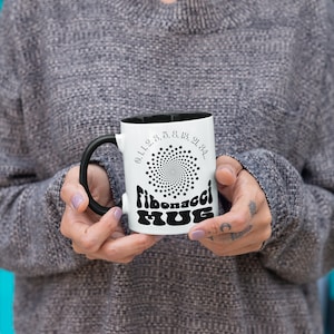 May include: White ceramic mug with a black handle. The mug has a black and white design with the text "Fibonacci Mug" and a spiral pattern of dots. The text "0, 1, 1, 2, 3, 5, 8, 13, 21, 34..." is printed around the spiral.