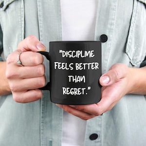May include: A black coffee mug with the quote "DISCIPLINE FEELS BETTER THAN REGRET." in white lettering. The mug is held by a person wearing a light green shirt and a white shirt underneath.