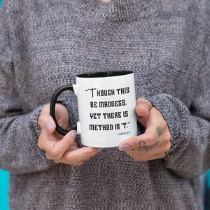 May include: White coffee mug with a black handle and black rim. The mug has a quote from Hamlet: "Though this be madness, yet there is method in 't."