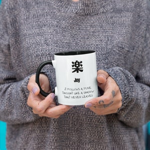 May include: White ceramic mug with a black handle. The mug has a black rim and a black kanji character on the front. The text "Joy" is printed below the character. The text "It follows a pure thought like a shadow that never leaves" is printed below the word "Joy".