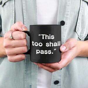 May include: A black coffee mug with the phrase "This too shall pass" in white lettering. The mug is held by a person wearing a light green shirt. The mug has a black handle.
